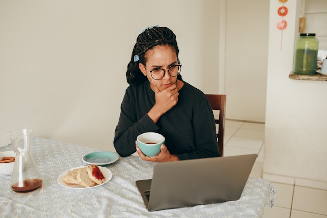 A woman holding a cup of tea looks confused as she stares at her laptop screen. 
