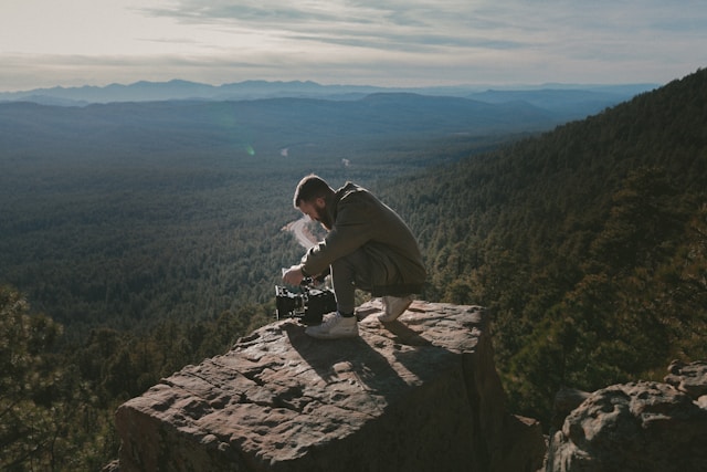 A man crouches on a rock ledge to capture a video of a mountainscape. 