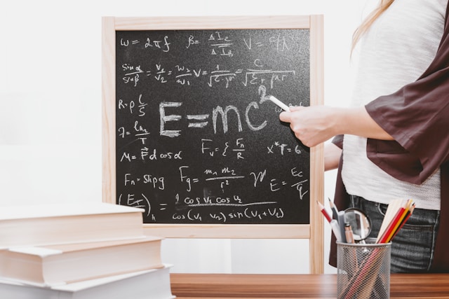 A teacher stands beside a small blackboard with various mathematical formulas on it. 
