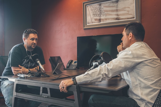 Two men sit face to face with microphones between them on a table. 