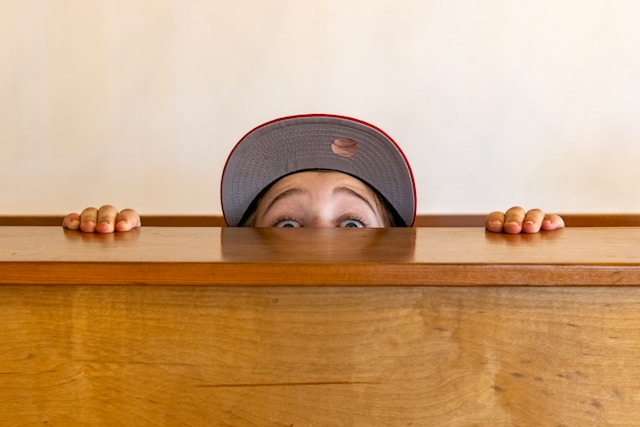 A person wearing a cap peeks over a wooden table. 
