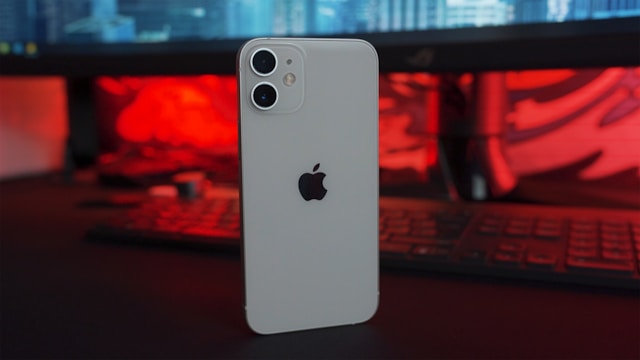 A white iPhone featuring the Apple logo on its back stands on a table in front of a desktop computer. 