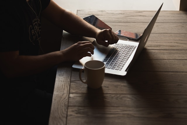 A person types on a laptop on a table beside a white coffee cup. 
