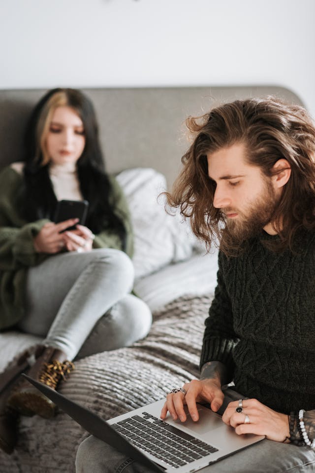 A woman uses her cell phone while a man uses his laptop.
