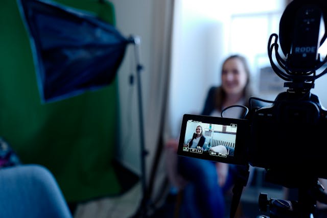 A black video camera records a woman by a window.
