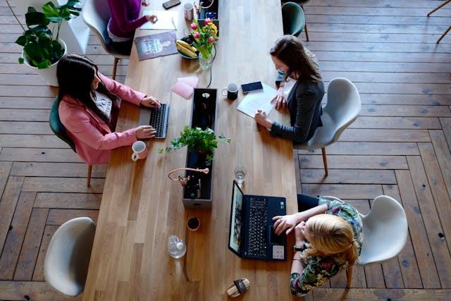 Three women do work at a wooden table. 
