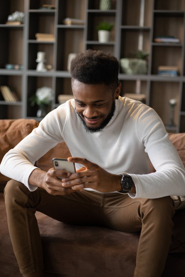 A man in a white sweater scrolls on his phone with a smile.
