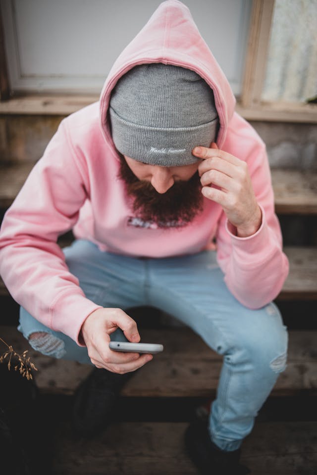 A man wearing a pink hoodie types on his cell phone.