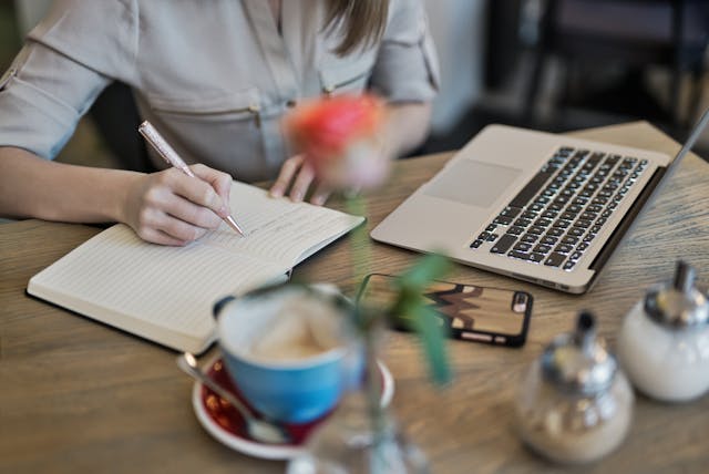 A person writes in a notebook next to a laptop.