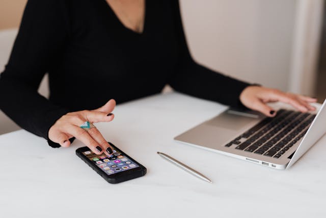 A woman scrolls on her phone while she uses her laptop.