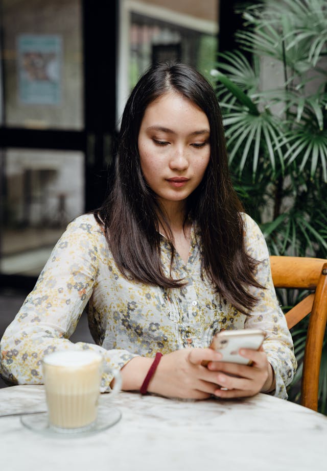 A woman types on her cell phone with a coffee on her table.