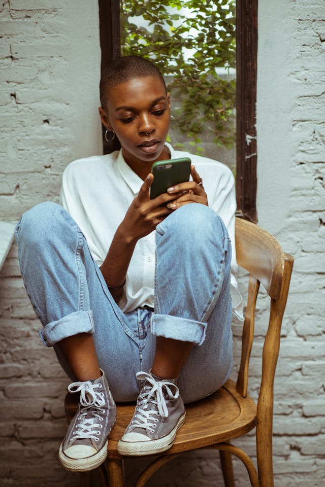 A woman sits on a wooden chair while scrolling on her phone.
