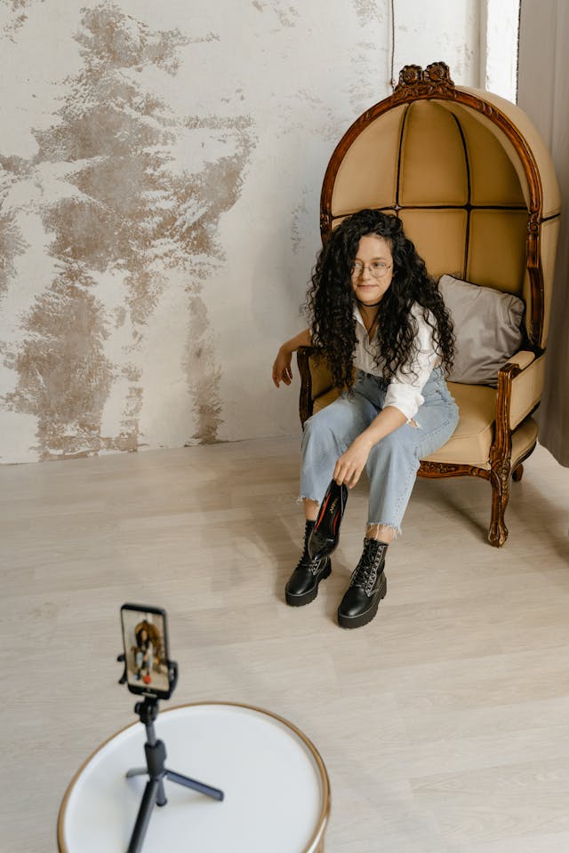 A woman records herself showing off a shoe as she sits in a large chair.
