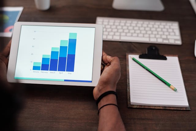 Someone holds a white iPad displaying a graph while a keyboard and book are on a table.
