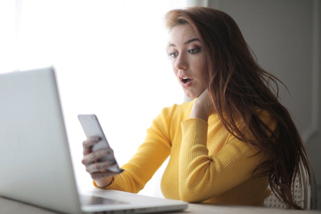 A woman in a yellow sweater looks at her phone with surprise.
