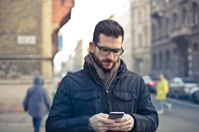 A man outside in a large jacket types on his phone.
