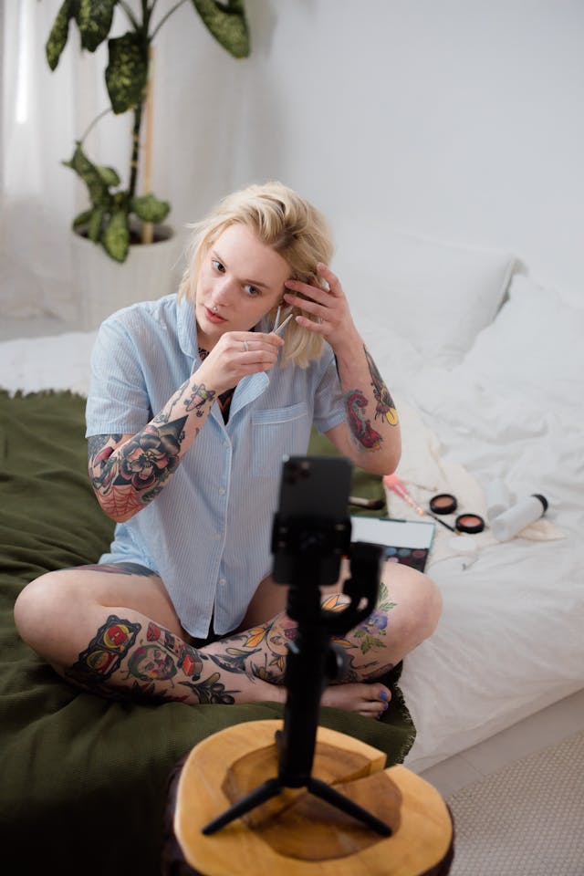 A woman records a makeup routine while she sits on her bed.
