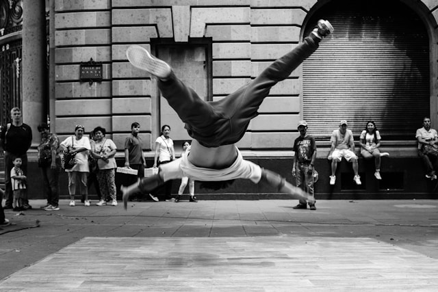 A male dancer executes a backflip in front of an audience in a busy street. 
