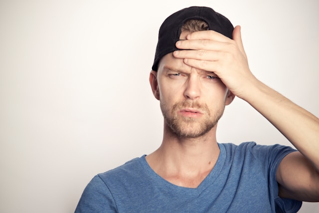 A man wearing a black cap backward looks confused and puts his hand on his forehead.