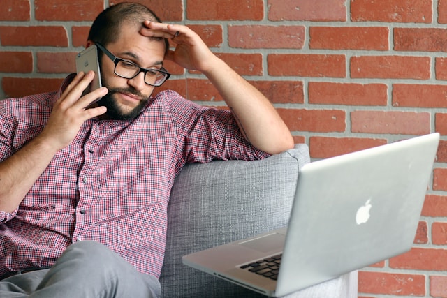 A man places a call with his iPhone while he sits and stares at his Macbook in frustration. 