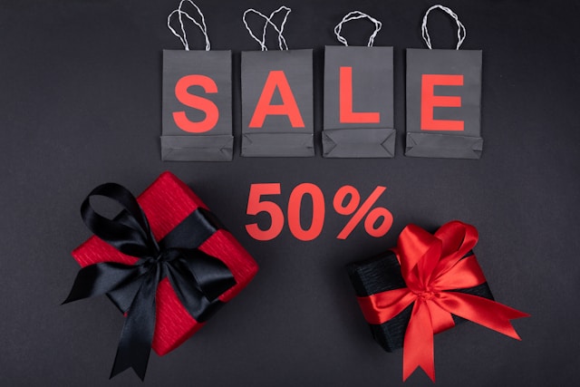 Four black shopping bags spell out the word “SALE,” and two gift boxes frame a 50% discount offer. 
