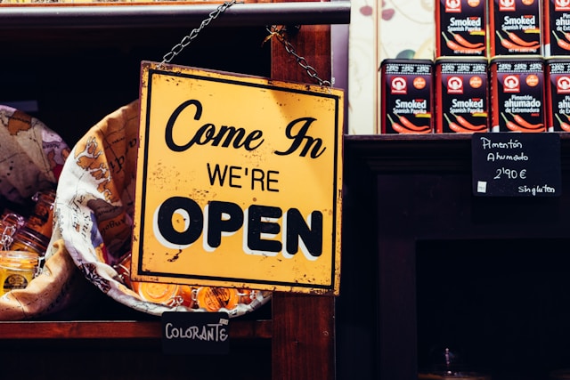 A black and yellow “Come in, we’re open” sign hangs in front of a store shelf. 
