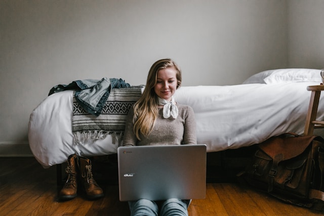 A woman sits on the floor in front of her bed as she browses TikTok on her laptop.