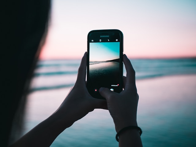 A person takes a photo of the beach at sunset using their phone to share on social media. 