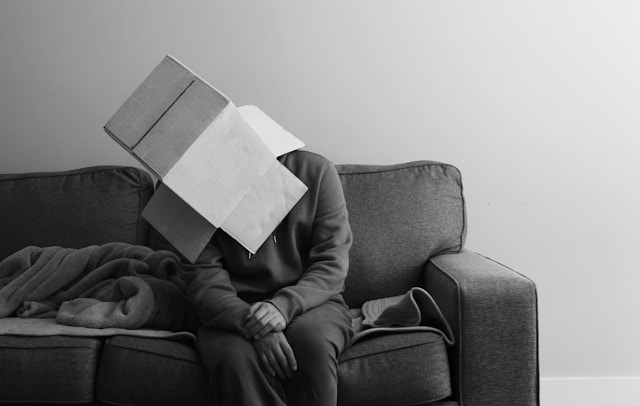 A person sitting on a couch wears a cardboard box on their head to hide their face. 
