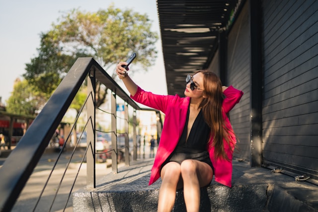 A woman in a pink blazer sits on concrete steps and takes a selfie for her TikTok page. 
