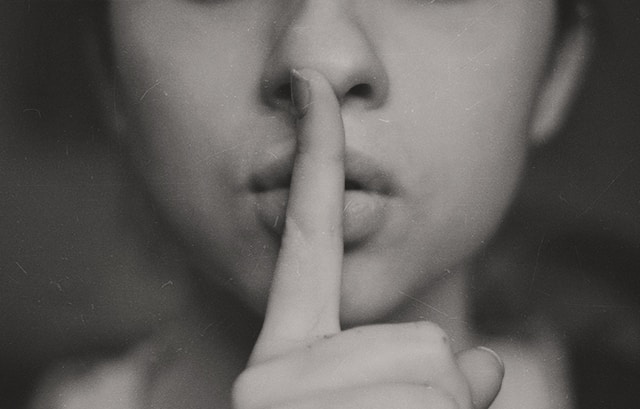 A woman makes the shushing sign with her index finger over her lips. 
