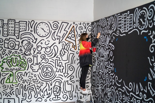 An artist on a step ladder draws doodles on a wall.