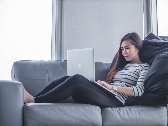 A woman lies on a couch and uses her laptop to watch TikTok videos. 
