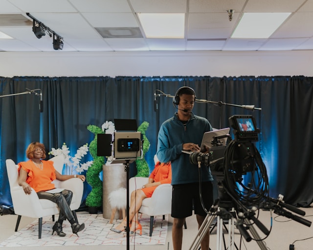 Two women sit on white chairs in front of cameras to host a live show. 
