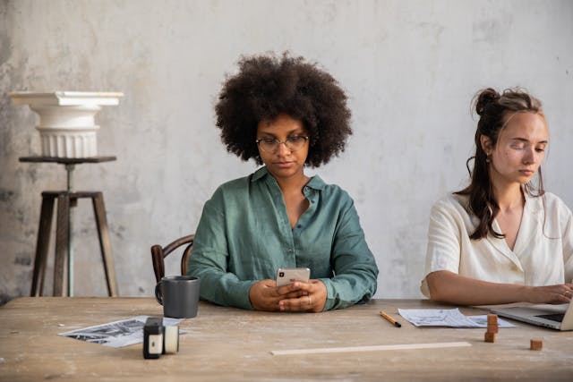 Two sit at a table typing on their devices.
