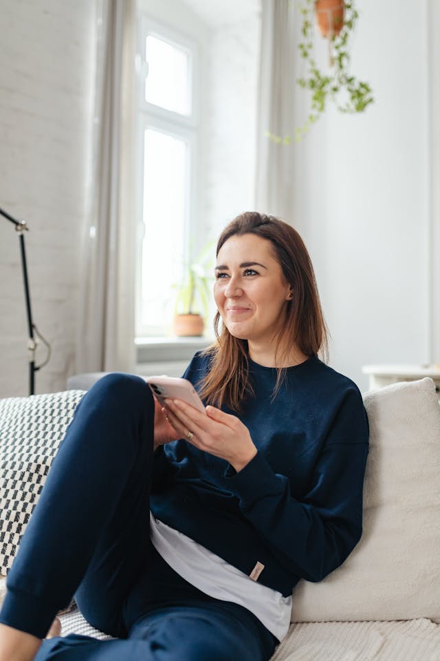 A woman smiles on the couch while she holds her phone.
