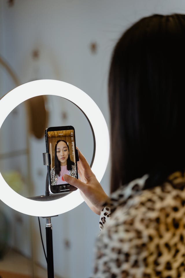 A woman clicks the record button on her cell phone mounted in front of a ring light.
