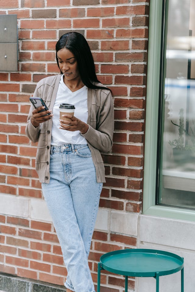 A woman looks at her phone screen and holds a cup of coffee.
