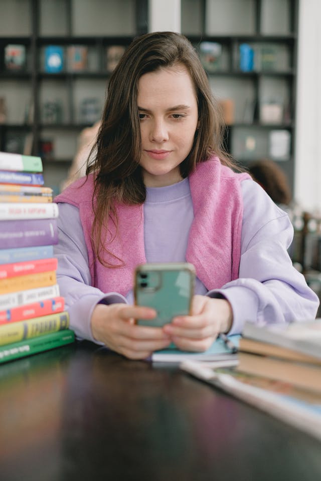 A woman uses a phone next to a stack of books.