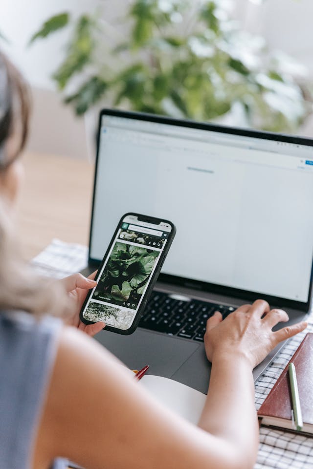 A person holds a phone in one hand and types on a laptop with the other hand.
