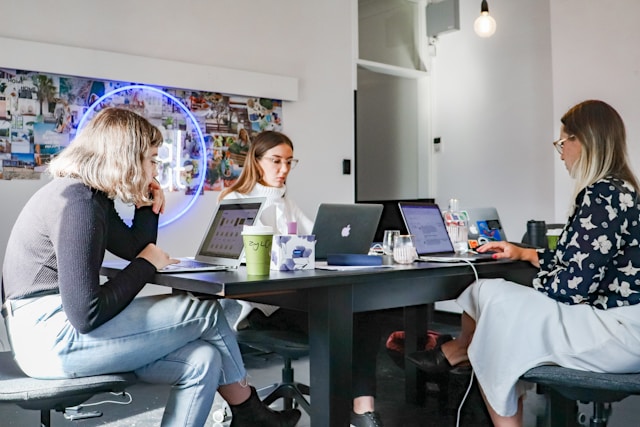 Three female professionals sit around a desk and work on their laptops. 