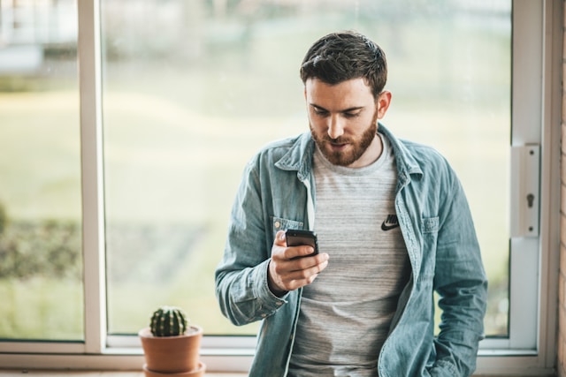 A man stands in front of a window and uses his phone to browse TikTok. 
