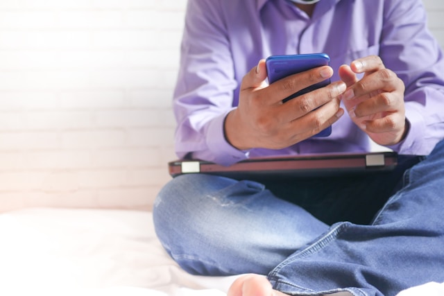 A person sits on the bed and browses on their phone. 
