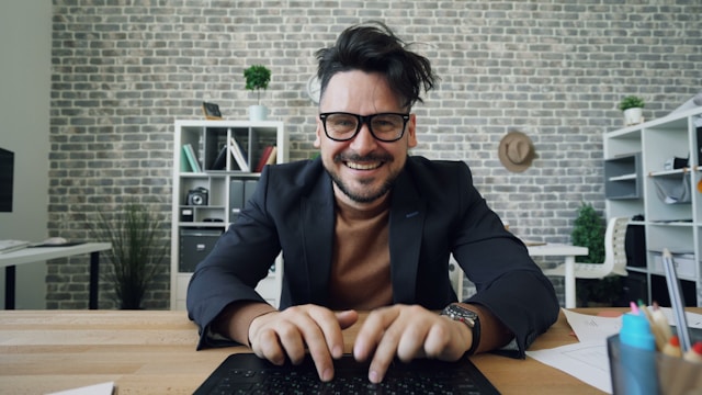 A man with glasses sits at a table and types comments on TikTok using his laptop. 
