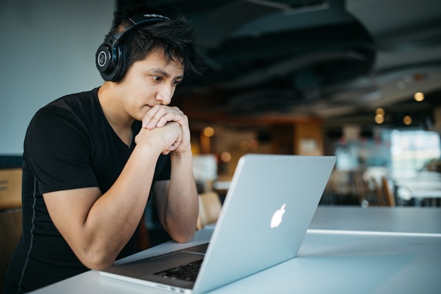 A man wearing headphones sits in a cafe and reads comments on his TikTok videos via his laptop. 
