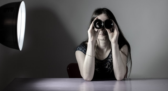 A woman sits on a table and uses a pair of binoculars. 
