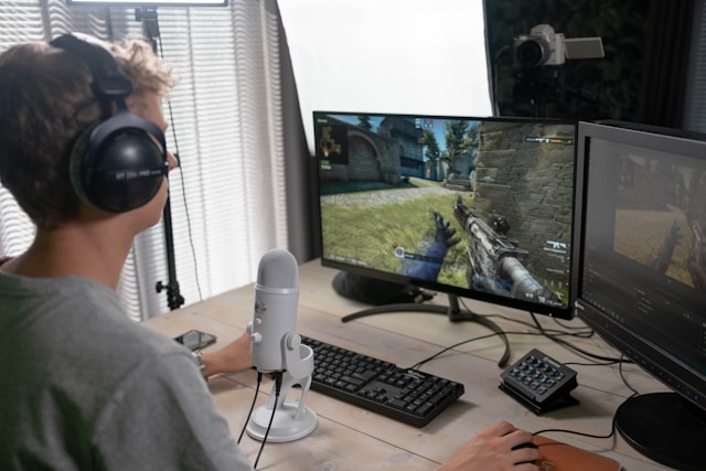 A video gamer sits in front of a camera, a microphone, and two computer monitors as he livestreams his game. 
