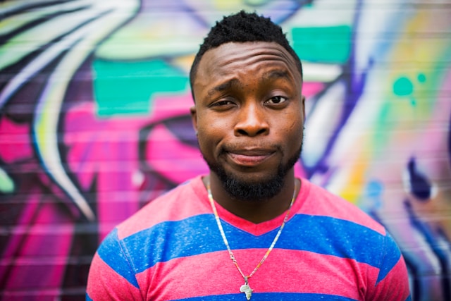 A man stands in front of a wall covered in graffiti and wears an expression of disbelief. 
