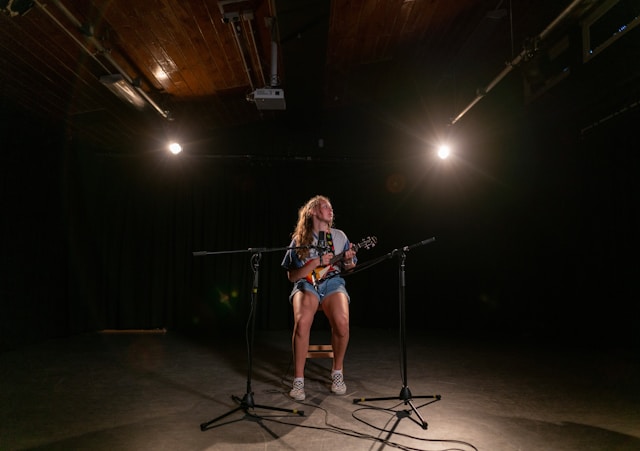 A musician plays the ukelele under two spotlights and in front of two microphones. 
