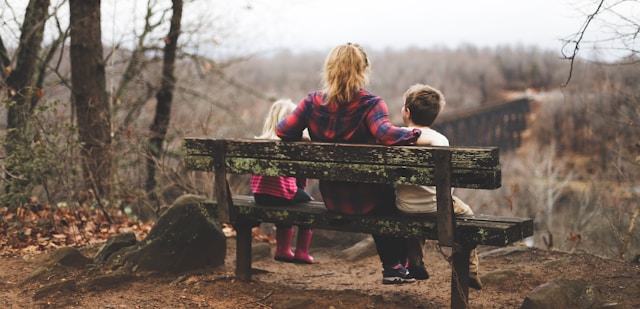 A mom sits between her kids on a wooden bench overlooking a bridge. 
